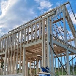 RKON Projekty i nadzór w budownictwie ROGALA KAMIL - Wzniesiony dwukondygnacyjny drewniany szkielet domu jednorodzinnego, z widocznymi pionowymi słupkami i poziomymi ryglami, na tle błękitnego nieba z białymi chmurami. Częściowo widoczna więźba dachowa.