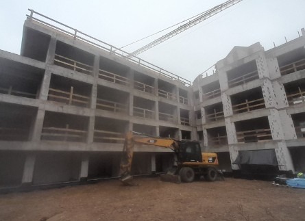 Surowy szkielet betonowy budynku wielopiętrowego w trakcie budowy, widoczny żółty koparko-ładowarka CAT i żuraw w tle, pochmurny dzień.