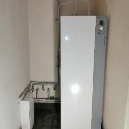 Biała pompa ciepła marki Atlantic z szarym panelem bocznym i rurami instalacyjnymi w rogu pomieszczenia z beżowymi ścianami i ciemną podłogą.