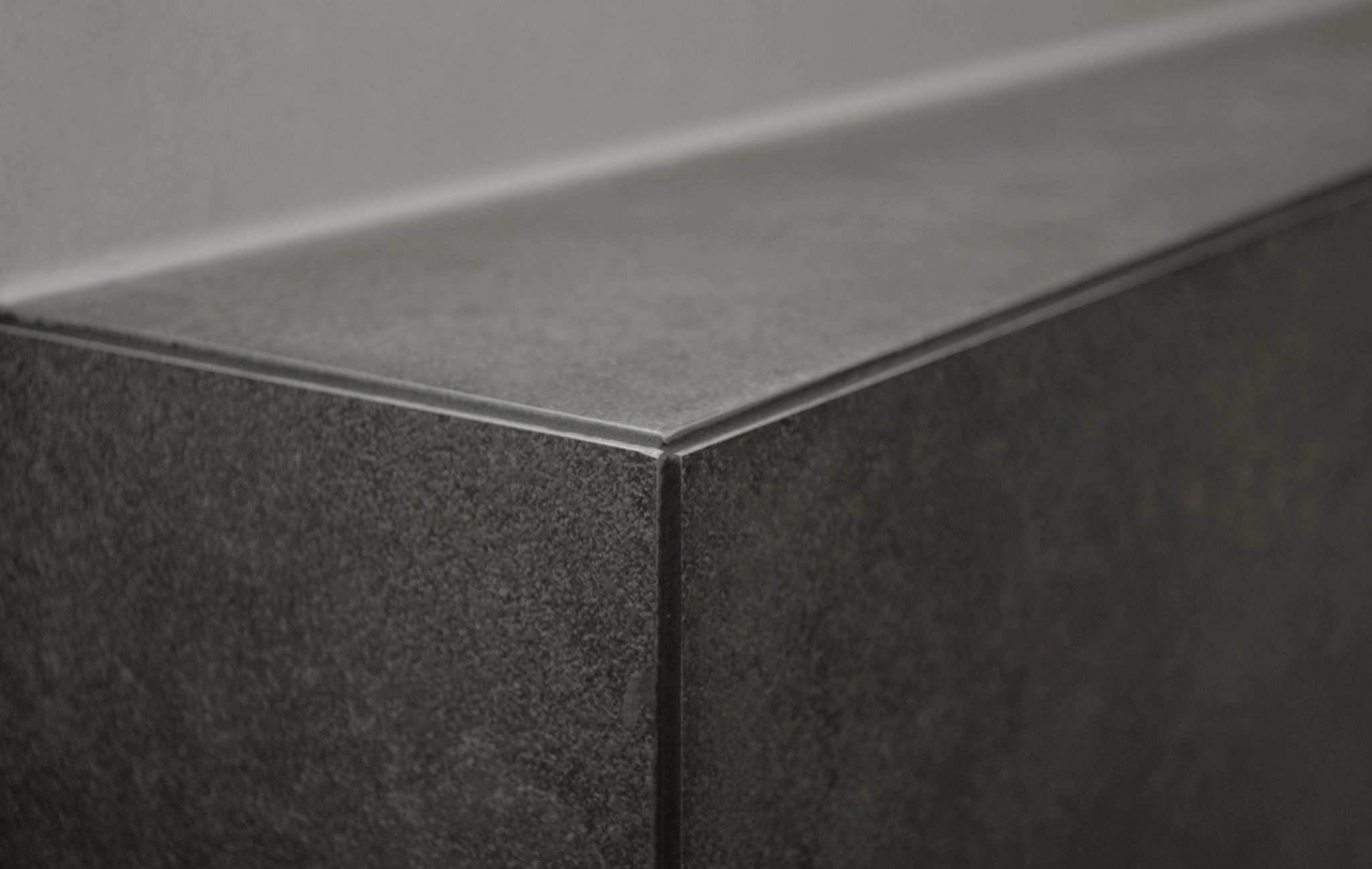 Szczegół narożnika blatu z ciemnoszarej, matowej płytki ceramicznej, wykończonego listwą, na tle jasnoszarej ściany.