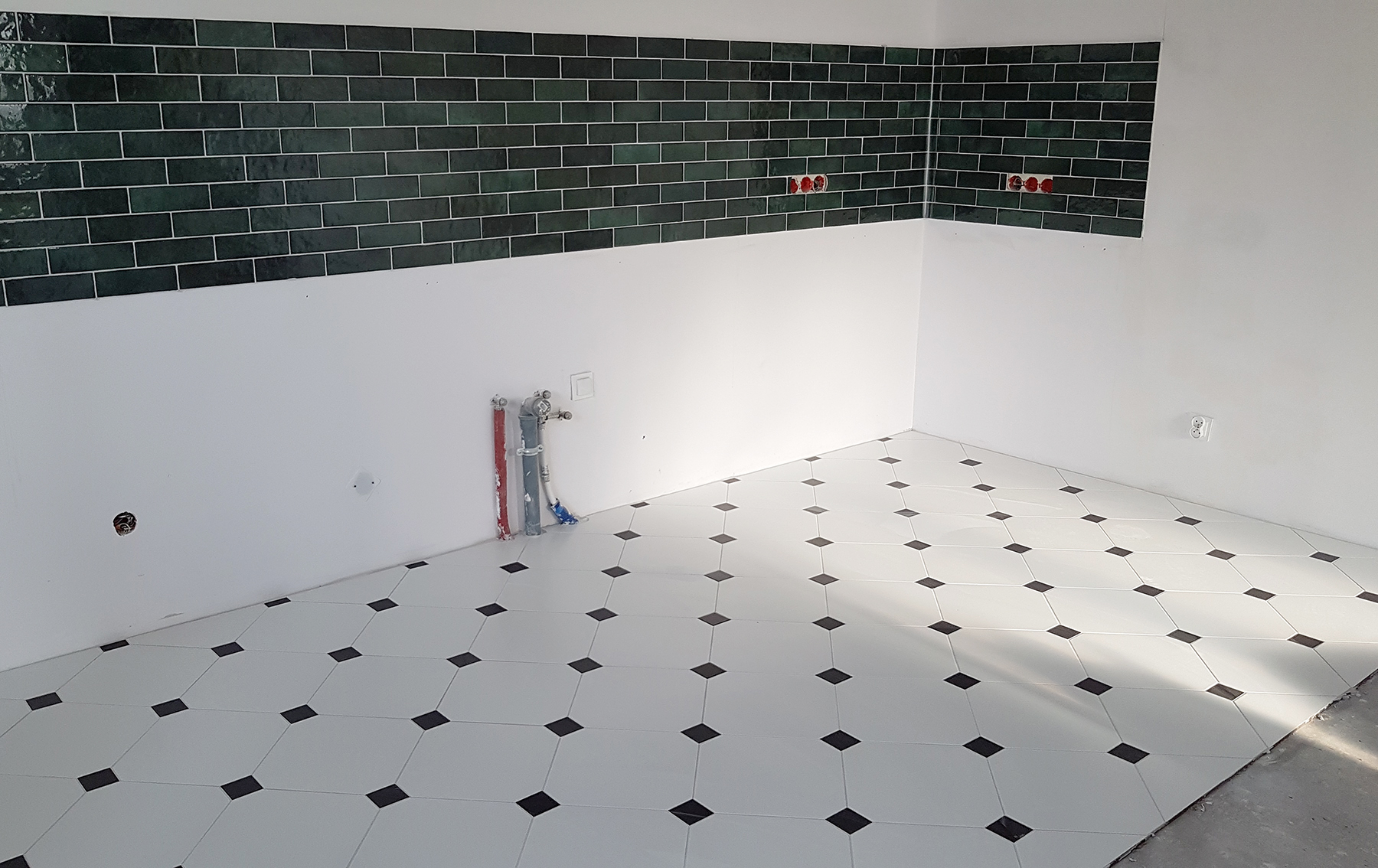 Wnętrze łazienki w trakcie remontu: ciemnozielona, cegiełkowa glazura na ścianie kontrastuje z białą farbą, a podłoga wyłożona jest biało-czarną płytką w romby. Widoczne instalacje wodne...