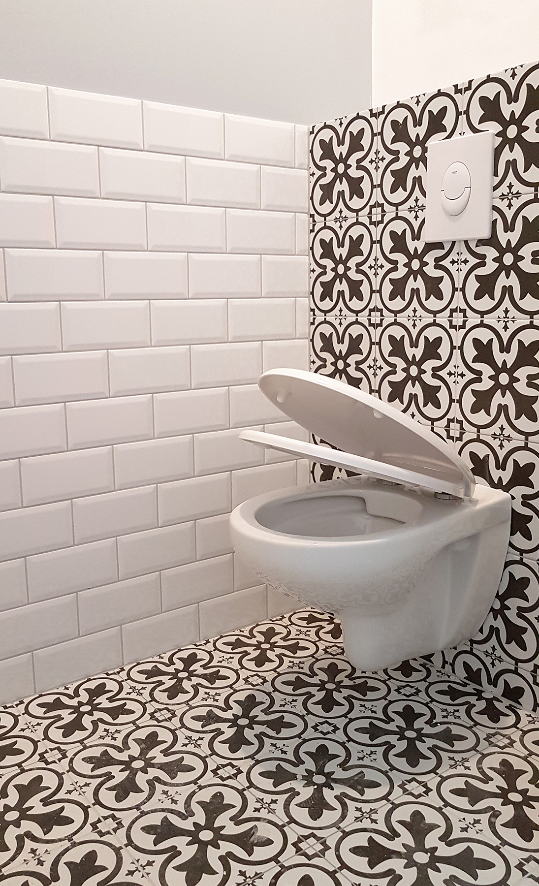 Nowoczesna toaleta podwieszana z otwartą deską, białe kafelki typu metro na ścianie, wzorzyste płytki podłogowe i ścienne w kolorze czarno-białym, przycisk spłuczki Grohe.