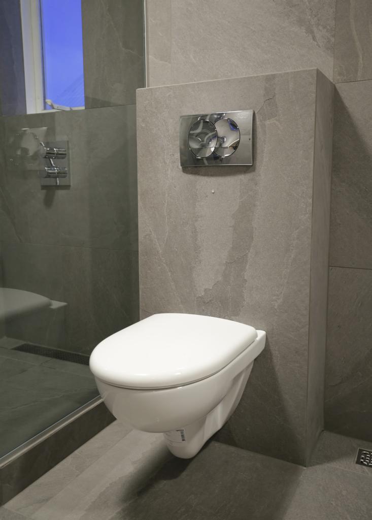 Podwieszana toaleta z chromowanym przyciskiem spłukującym w łazience wykończonej szarymi płytkami imitującymi kamień.