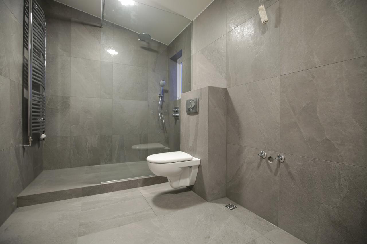 Nowoczesna łazienka wykończona szarymi płytkami imitującymi kamień, z kabiną prysznicową typu walk-in i podwieszaną toaletą.