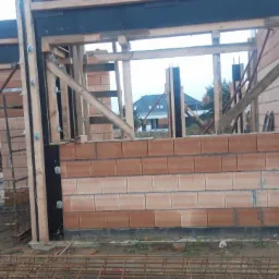 Wznoszenie muru z czerwonej cegły ceramicznej w trakcie budowy domu. Widoczne drewniane szalunki okienne i stalowe elementy konstrukcyjne. W tle zarys budynku mieszkalnego.