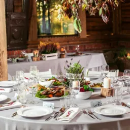 Elegancko nakryty okrągły stół z szarą pościelą, białymi talerzami, srebrnymi sztućcami i dekoracjami roślinnymi, ustawiony w rustykalnej sali z drewnianymi elementami i ceglanymi ścianami...