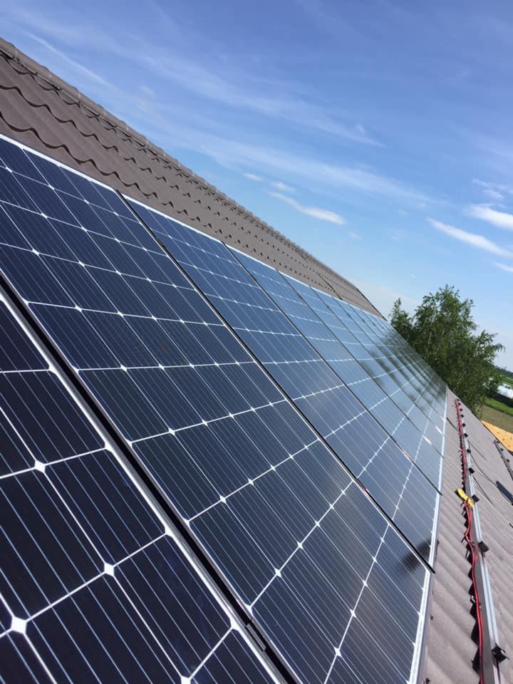 Panele słoneczne zainstalowane na brązowym dachu z dachówki ceramicznej, widoczne przewody i elementy montażowe, błękitne niebo w tle.