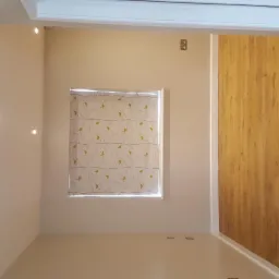 Wnętrze po remoncie: gładkie, beżowe ściany z punktowym oświetleniem, roleta w oknie z motywem gałązek, oraz panelowa ściana z drewnopodobnego materiału.