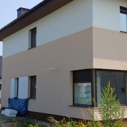 Adrian marek - Dwukondygnacyjny dom z częściowo ocieploną elewacją. Widoczne białe płyty izolacyjne przy oknie, przygotowane do montażu. Na pierwszym planie pracownicy i materiały budowlane.