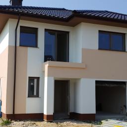 Adrian marek - Dwukondygnacyjny dom w trakcie prac wykończeniowych elewacji, z widocznym pracownikiem na rusztowaniu malującym ścianę na jasny kolor, w tle drugi dom z ciemnym dachem.