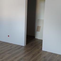 Adrian marek - Wnętrze pokoju po remoncie, z nową podłogą z paneli imitujących drewno i świeżo pomalowanymi na biało ścianami z widocznym otworem drzwiowym prowadzącym do kolejnego pomieszczenia, w którym oparta...