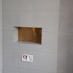 Adrian marek - Ściana łazienki z szarymi płytkami, wnęka z mozaiką w odcieniach złota, zabudowany stelaż podtynkowy WC oraz przyłącza kanalizacyjne w podłodze, widoczna izolacja w rogu.