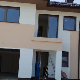 Adrian marek - Dwukondygnacyjny dom jednorodzinny w trakcie prac wykończeniowych elewacji, widoczne fragmenty ocieplenia i świeżo pomalowane fragmenty w kolorze beżowym i białym, w tle pracownik bez koszulki.