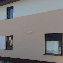 Adrian marek - Pracownik w kombinezonie maluje fragment elewacji dwukolorowego domu jednorodzinnego na biało, stojąc na prowizorycznym podeście z bloczków, widoczne puszki z farbą, w tle fragment ogrodu i samochód.