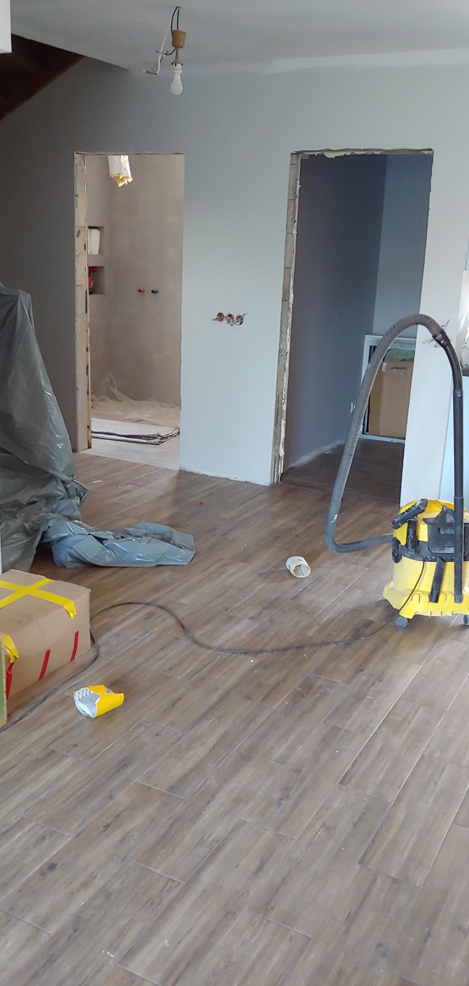 Wnętrze w trakcie prac wykończeniowych: widoczne niezabezpieczone otwory drzwiowe, podłoga wyłożona panelami, resztki materiałów budowlanych i odkurzacz przemysłowy.