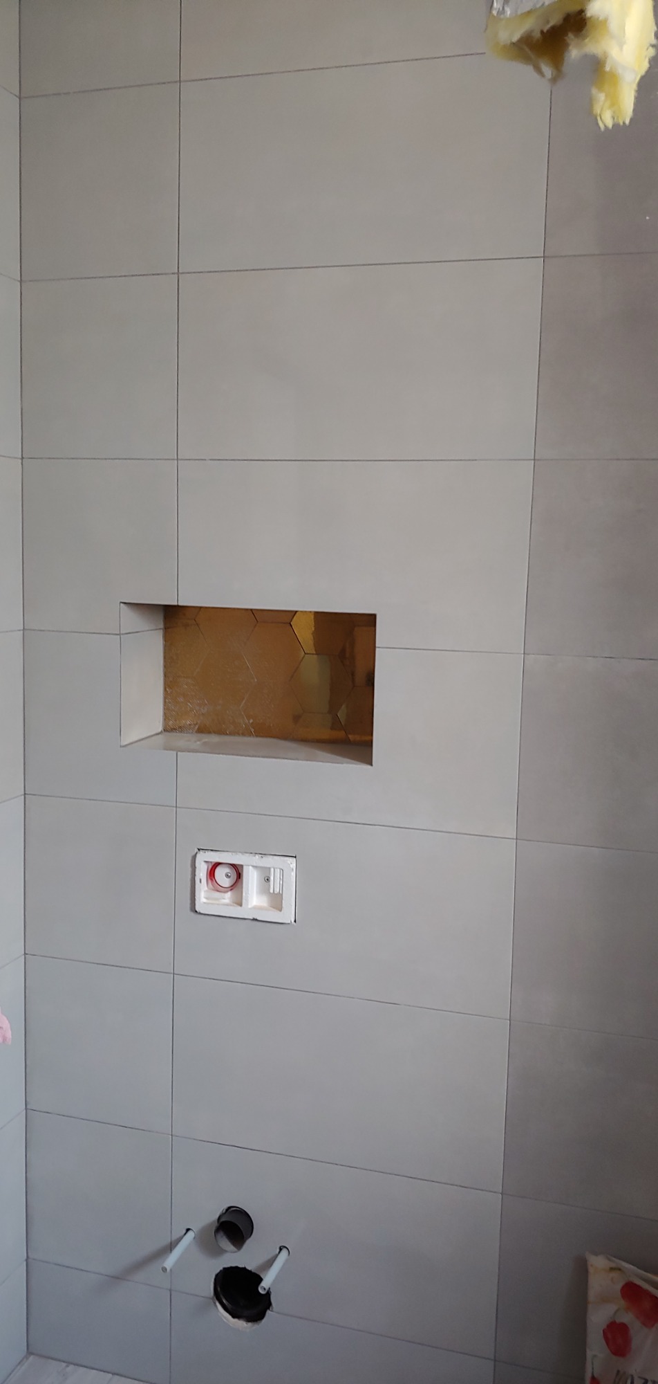 Ściana łazienki z szarymi płytkami, wnęka z mozaiką w odcieniach złota, zabudowany stelaż podtynkowy WC oraz przyłącza kanalizacyjne w podłodze, widoczna izolacja w rogu.