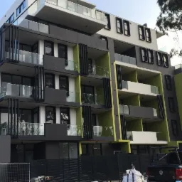 Nowoczesny budynek wielorodzinny z balkonami w odcieniach szarości, bieli i limonki, widoczny z poziomu gruntu, z fragmentem czarnego pickupa w dolnym rogu.