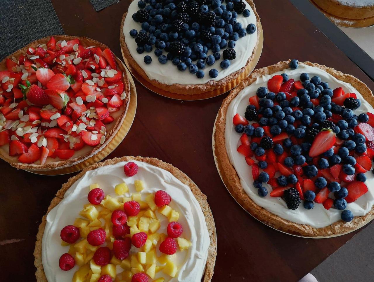 Cztery tarty na drewnianym stole: truskawkowa z migdałami, borówkowa, mieszana z truskawkami i borówkami oraz z malinami i mango.