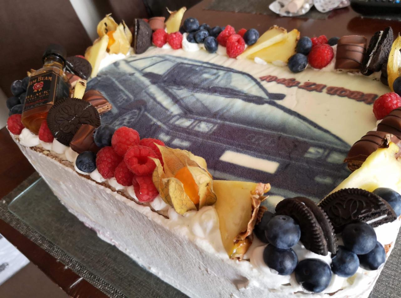 Prostokątny tort urodzinowy z nadrukiem samochodu, dekorowany bitą śmietaną, malinami, borówkami, physalis, kawałkami czekolady i buteleczką whisky Jim Beam.