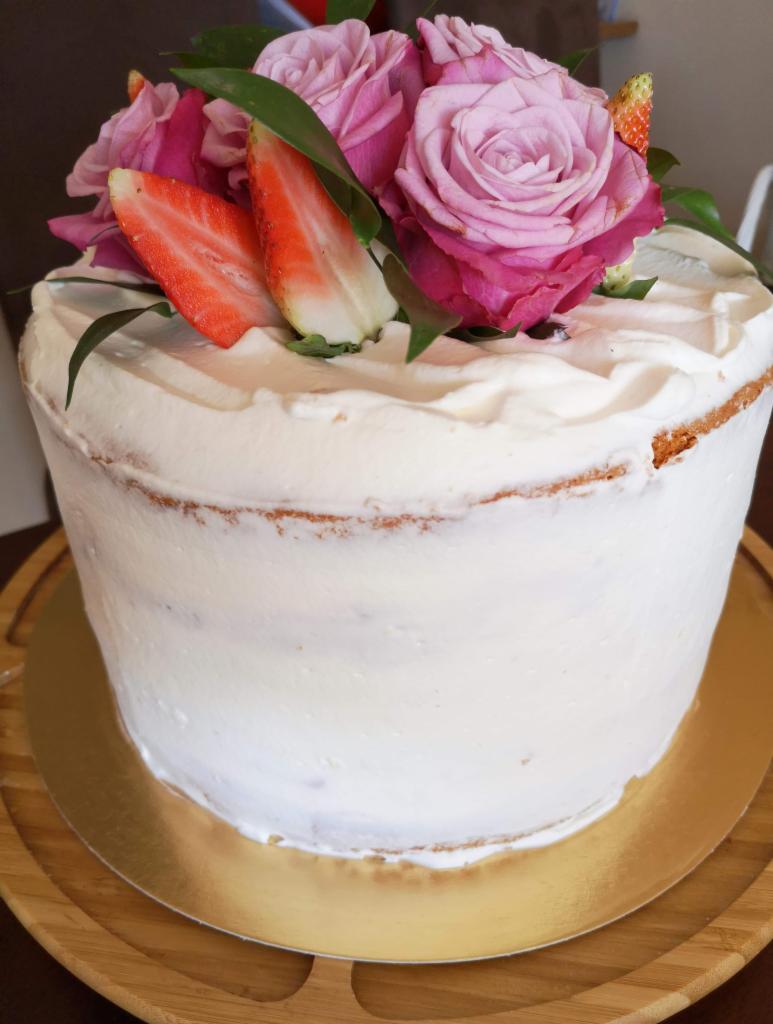 Tort bezowy z kremem i dekoracją z różowych róż oraz świeżych truskawek na drewnianej tacy.