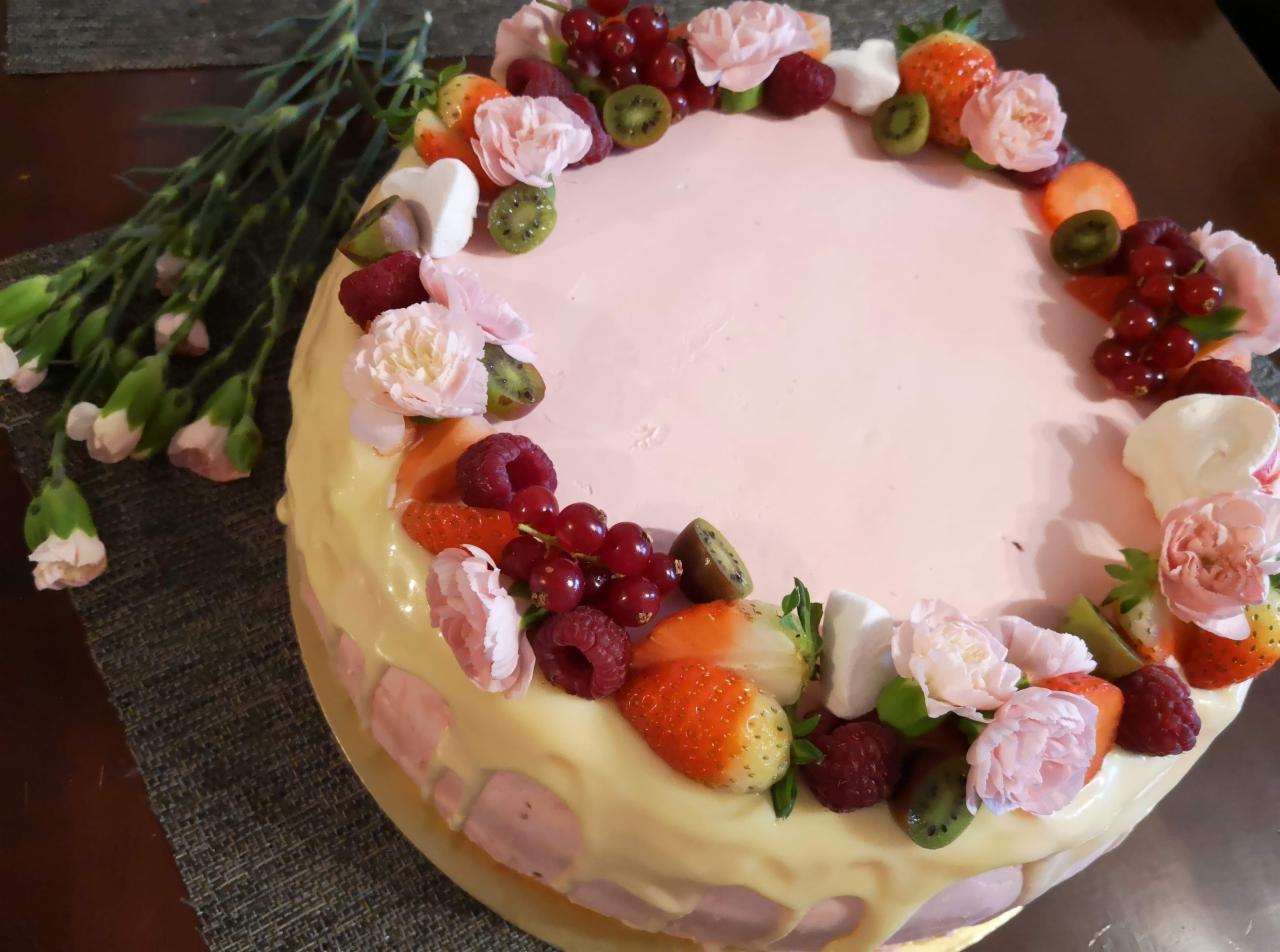 Tort urodzinowy z różowym kremem, polany białą polewą, udekorowany świeżymi truskawkami, malinami, porzeczkami, kiwi, piankami i różowymi goździkami.