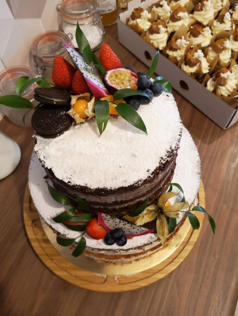 Dwupiętrowy tort bezowy z kremem, udekorowany świeżymi owocami: truskawkami, smoczym owocem, borówkami, physalis i ciasteczkami Oreo, na drewnianej paterze, w tle pudełko z babeczkami.