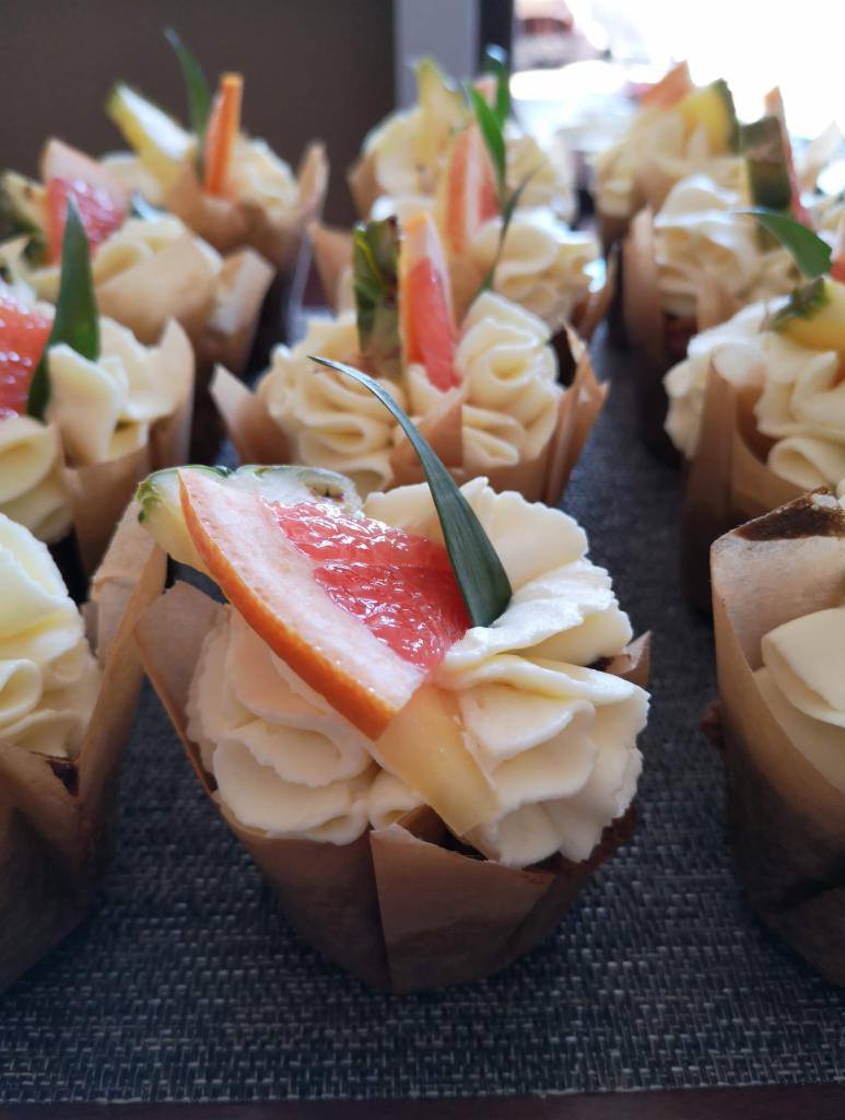 Mini babeczki z kremem i dekoracją z grejpfruta, ananasa i liścia, podane w papierowych foremkach na ciemnym tle.