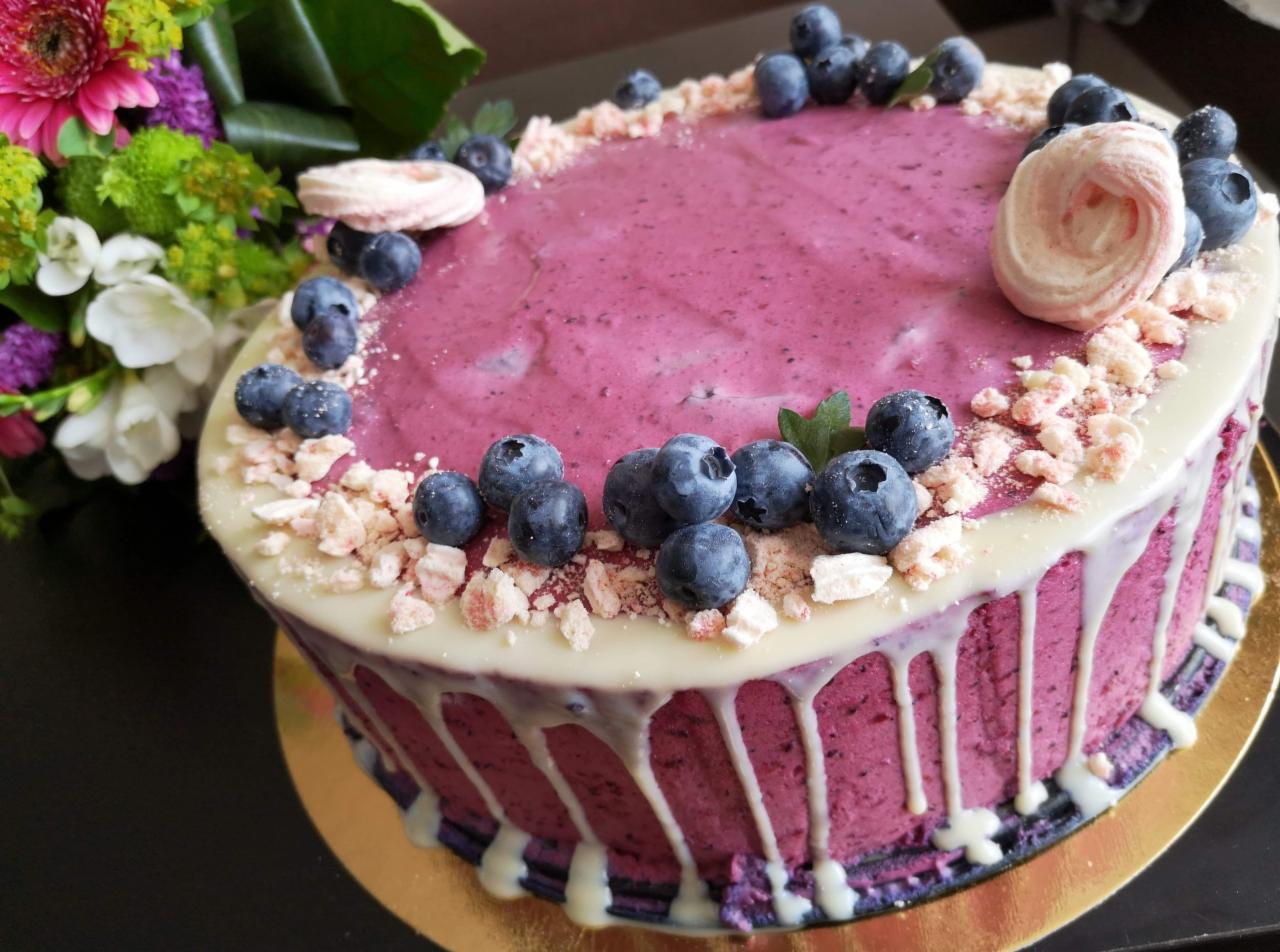 Tort borówkowy z białą polewą, posypany kruszonką i udekorowany świeżymi borówkami oraz bezą w kształcie róży, na złotym podkładzie, z bukietem kwiatów w tle.