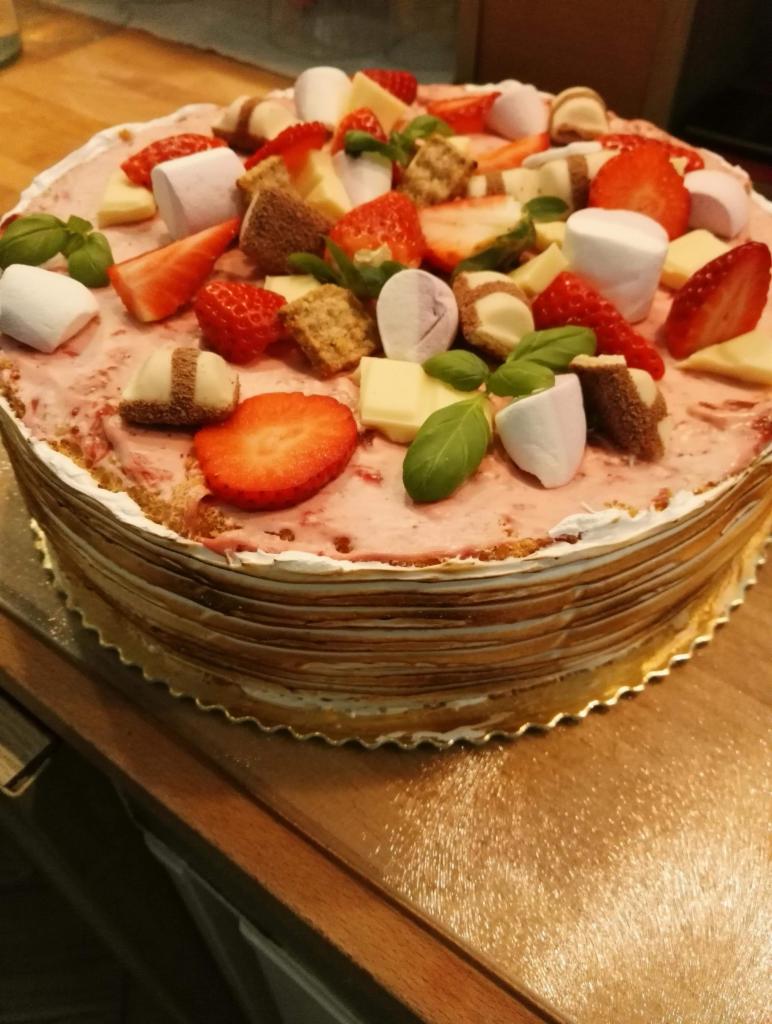 Wielowarstwowy tort z kremem, udekorowany truskawkami, piankami marshmallow, kawałkami białej czekolady, kostkami ciasta i listkami bazylii na złotej podkładce.
