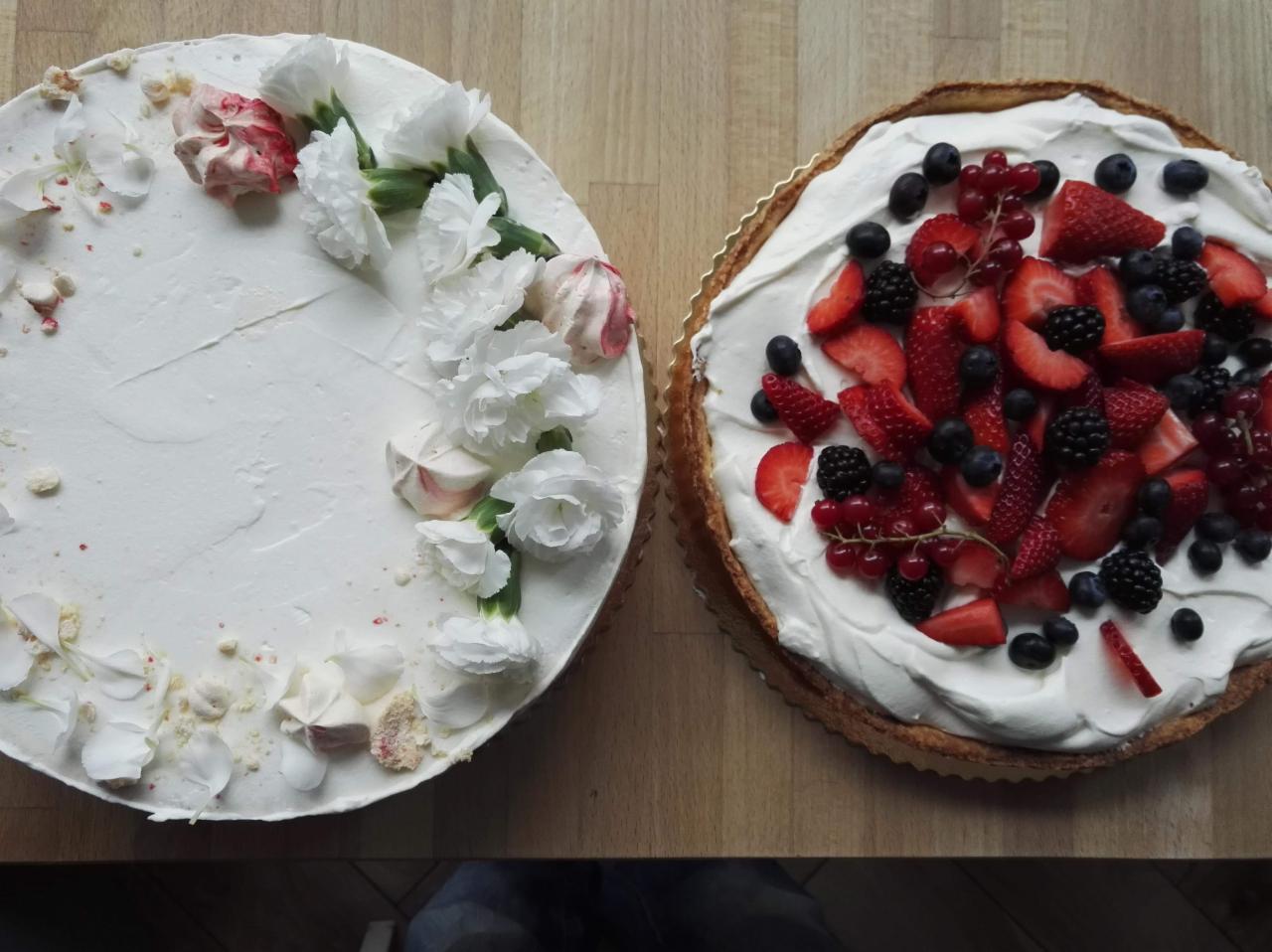 Dwa okrągłe torty: jeden z białym kremem i dekoracją z białych kwiatów oraz różowych bezików, drugi z kremem i mieszanką świeżych owoców: truskawek, borówek, jeżyn i czerwonych porzeczek,...