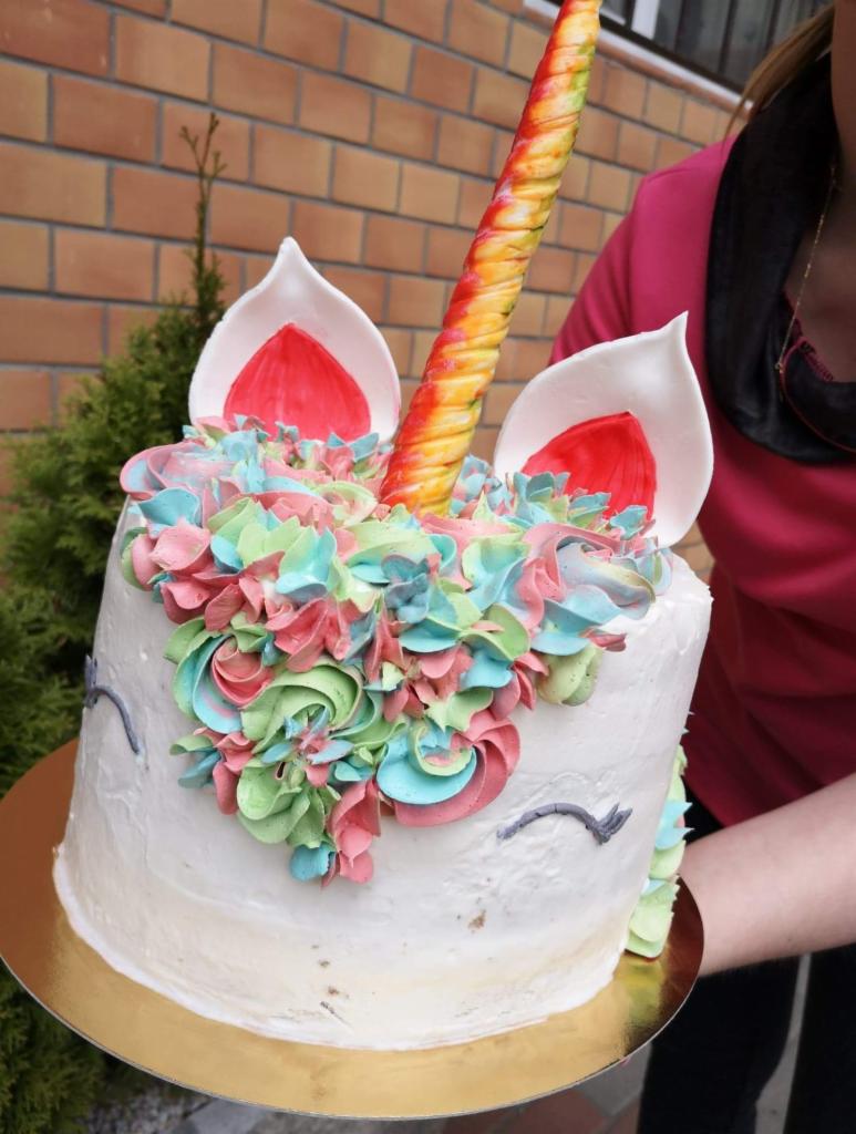 Tort jednorożec z kolorową grzywą z kremu, złotym rogiem i uszami, trzymany przez osobę na złotym podkładzie, na tle ceglanej ściany i krzewu.