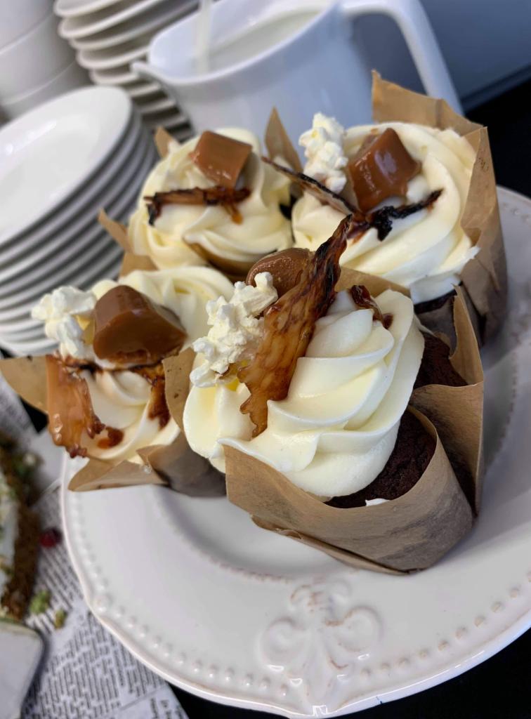 Cztery babeczki w papierowych foremkach na białym talerzu, udekorowane kremem, karmelem i kandyzowaną skórką pomarańczową, w tle stos naczyń i dzbanek.