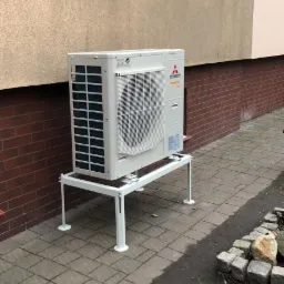 Biała jednostka zewnętrzna klimatyzacji Mitsubishi ustawiona na metalowym stelażu przy ścianie budynku z ceglanym cokołem i oknem na górze.