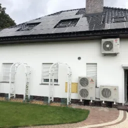 Biały dom z panelami słonecznymi na dachu i klimatyzacją Mitsubishi na ścianie, widoczne białe pergole w ogrodzie.