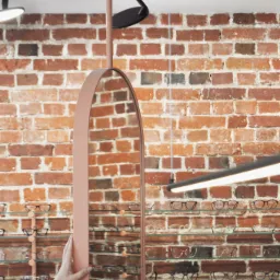 Owalne lustro w pudrowo-różowej ramie, ustawione w salonie optycznym na tle ściany z czerwonej cegły i szklanych półek z okularami.