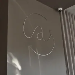 Metalowe logo firmy na szarej ścianie, obok zasłon, z widoczną kamerą monitoringu na suficie