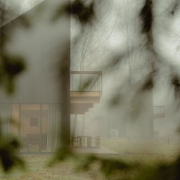 Madde studio Sp. z o.o. - Nowoczesny dom z drewnianym tarasem i minimalistycznymi meblami ogrodowymi, otoczony mglistym lasem, widziany przez gałęzie drzewa.