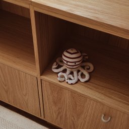 Madde studio Sp. z o.o. - Drewniany regał z szufladami i półkami, na których leży pluszowa zabawka ośmiornica w paski. Minimalistyczny design, ciepłe oświetlenie, dywan w tle.