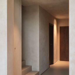 Madde studio Sp. z o.o. - Korytarz w odcieniach beżu z minimalistycznymi schodami i oświetleniem punktowym. Widoczne dwie drewniane drzwi i gładkie ściany.