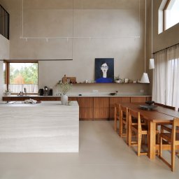 Madde studio Sp. z o.o. - Jasna kuchnia z wyspą z trawertynu, drewnianymi szafkami i stołem. Obraz z kobietą na ścianie. Minimalistyczne lampy wiszące i wysokie okno z widokiem na zieleń.