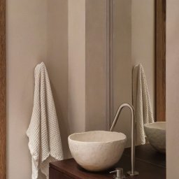 Madde studio Sp. z o.o. - Minimalistyczna łazienka: kremowa ściana z ręcznikiem, umywalka w kształcie misy na ciemnym blacie, nowoczesna bateria i lustro z drewnianą ramą.