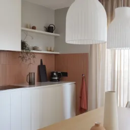 Minimalistyczna kuchnia w neutralnych barwach: białe szafki, terakotowa mozaika na ścianie, drewniany blat stołu z dekoracyjnymi wazonami i szklanką, wiszące białe lampy z plisowanym abażurem.