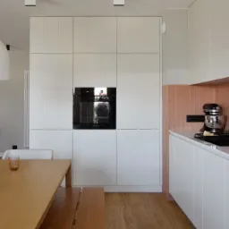 Jasna kuchnia z zabudową meblową sięgającą sufitu, drewnianym stołem z ławką i akcentem w postaci różowych płytek nad blatem.