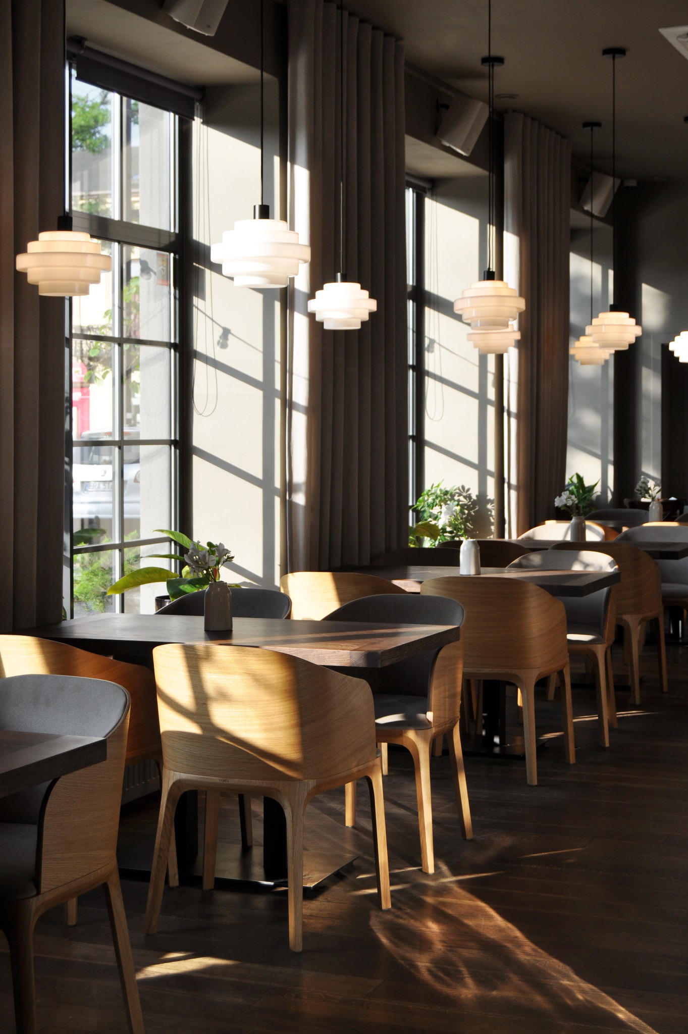 Jasne, nowoczesne wnętrze restauracji z drewnianymi krzesłami, ciemnymi stołami i wiszącymi lampami w stylu skandynawskim, oświetlone popołudniowym słońcem wpadającym przez okna.