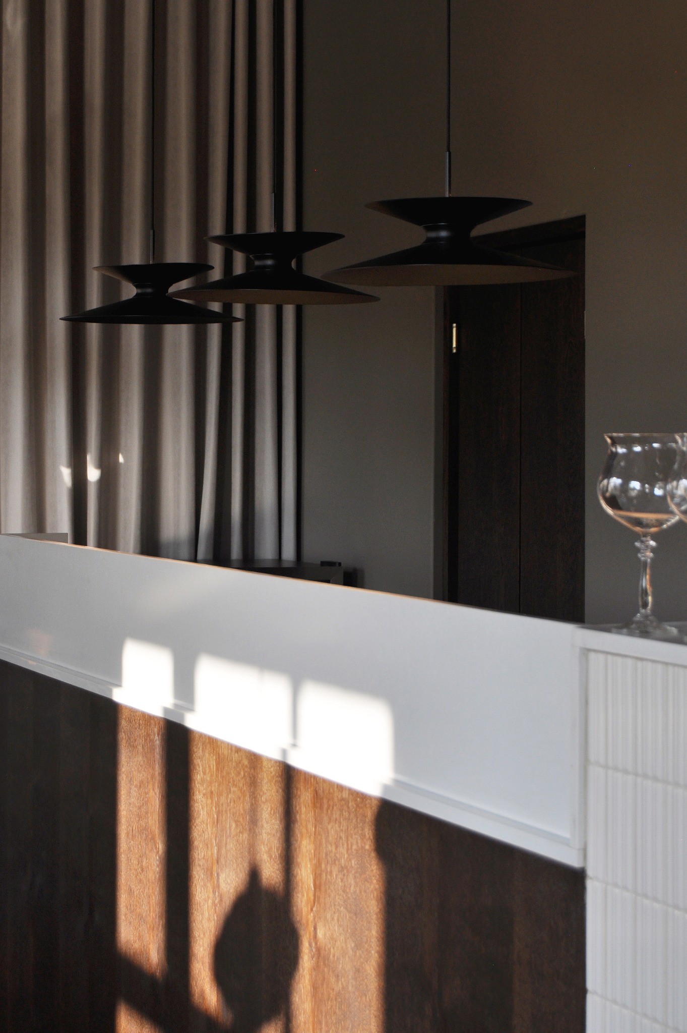 Nowoczesne lampy wiszące z czarnymi abażurami nad drewnianą zabudową z białym blatem, w tle zasłony i drzwi, minimalistyczny wystrój wnętrza.