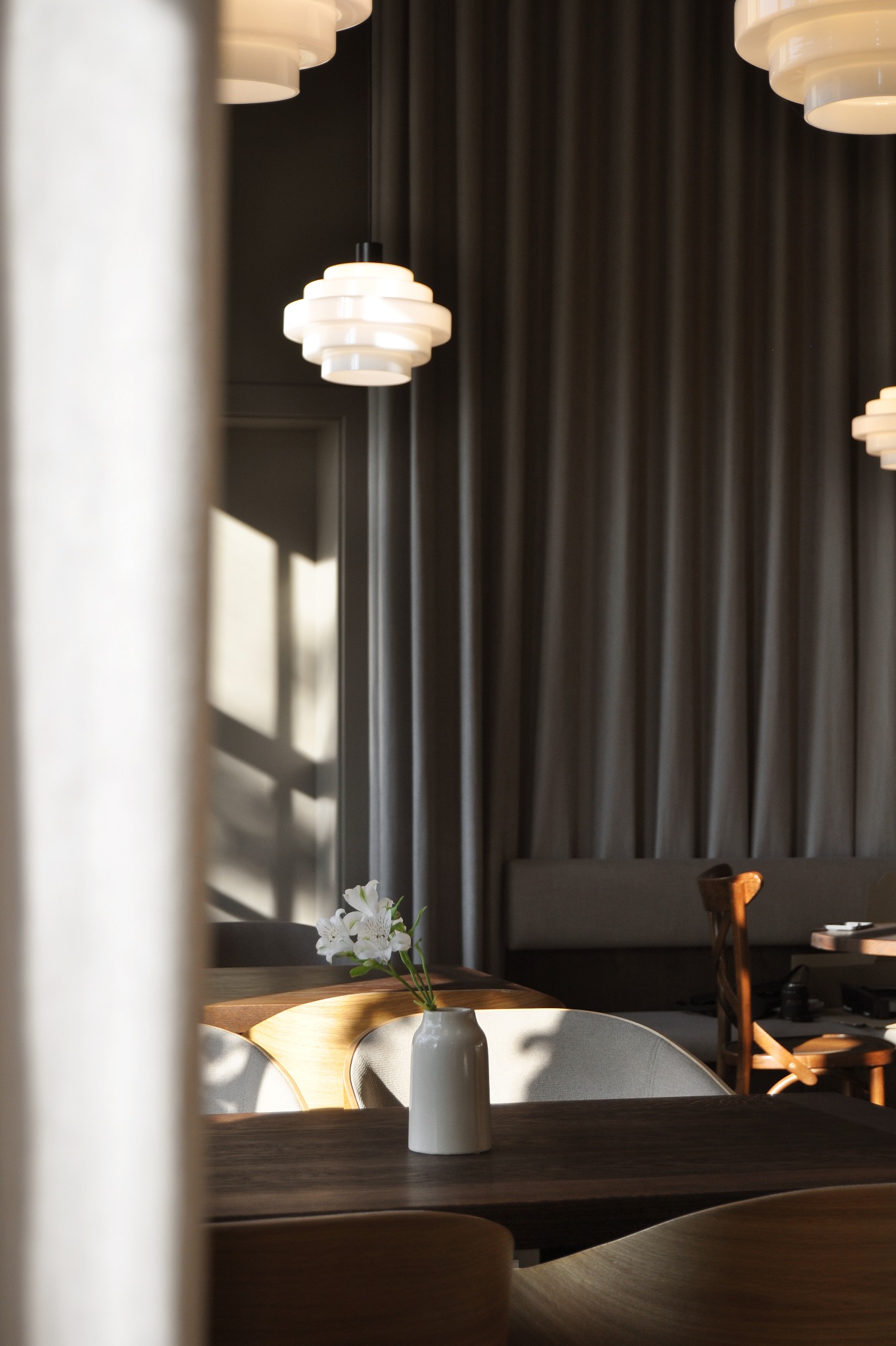 Minimalistyczna aranżacja wnętrza restauracji: białe kwiaty w ceramicznym wazonie na ciemnym drewnianym stole, nowoczesne lampy wiszące i zasłony w tle, widok zza białej kolumny.