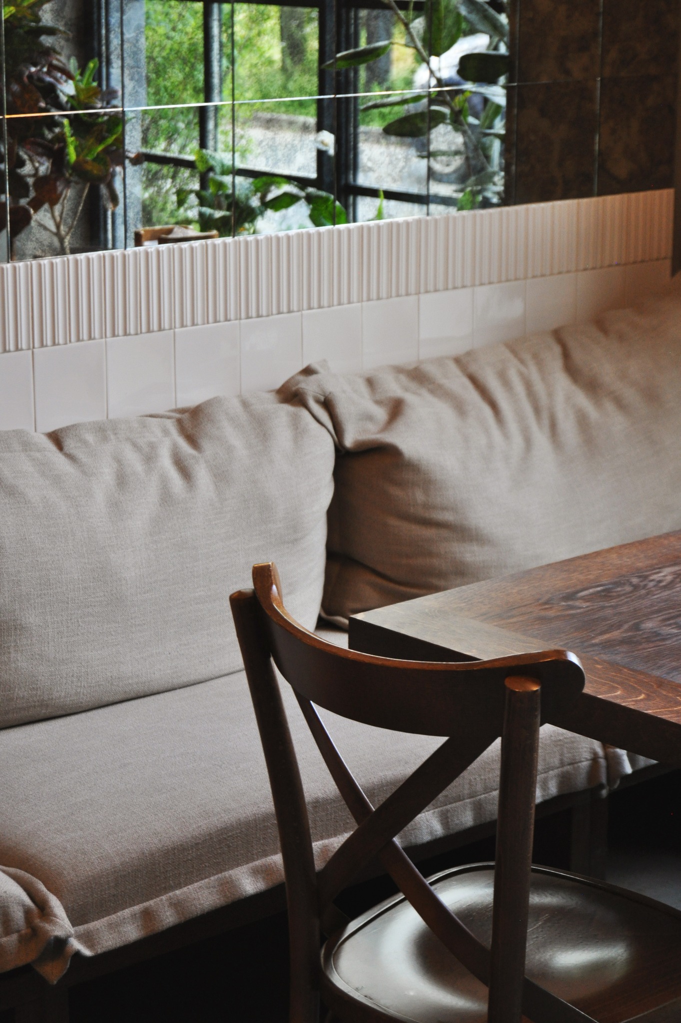 Beżowa tapicerowana ławka pod oknem z widokiem na zieleń, drewniany stół i krzesło w stylu skandynawskim, białe płytki na ścianie.