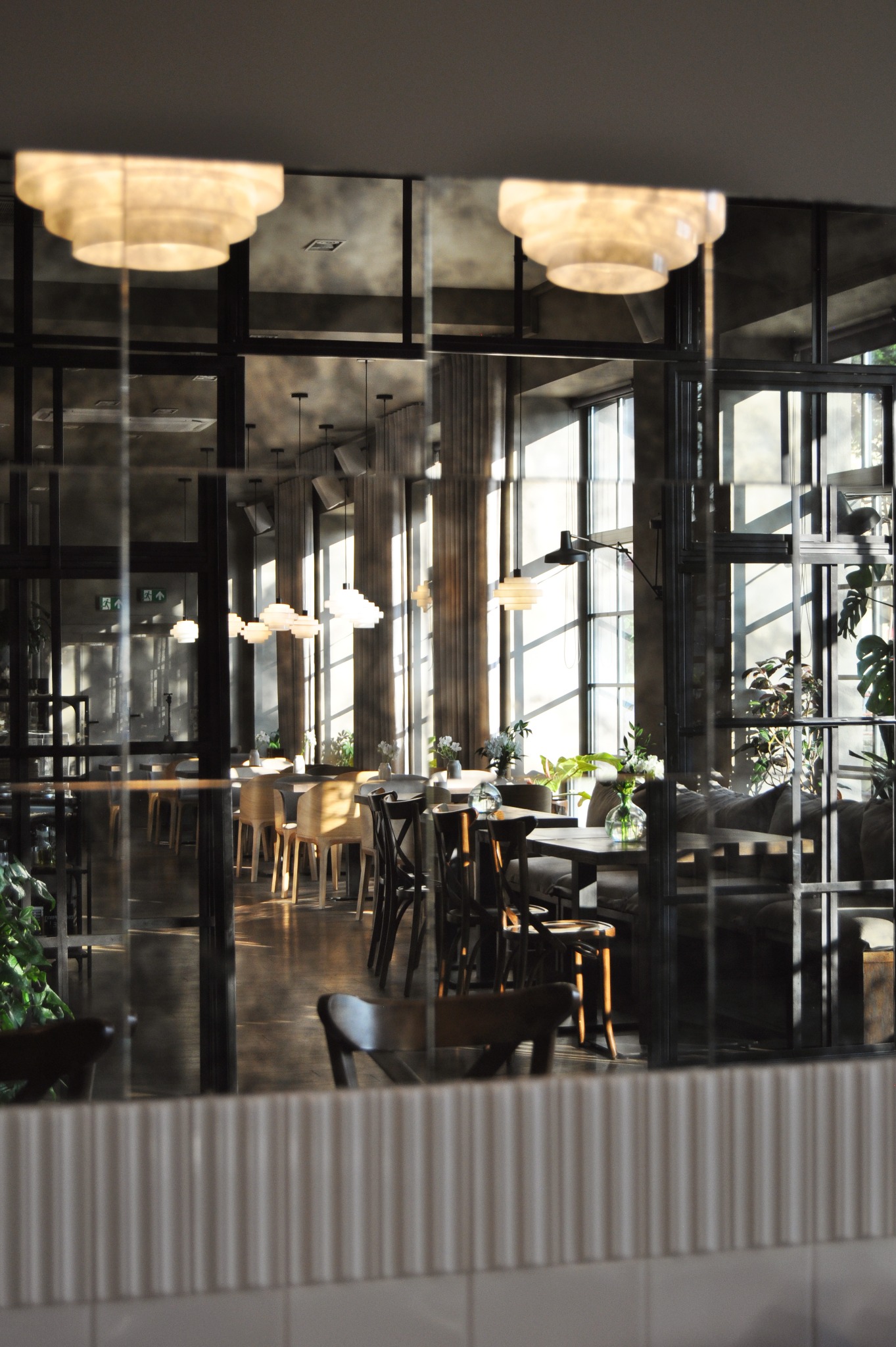 Eklektyczne wnętrze restauracji widziane przez lustrzaną ścianę, ukazujące drewniane krzesła, stoliki z bukietami kwiatów i wiszące lampy w stylu art deco, oświetlone naturalnym światłem wpadającym...