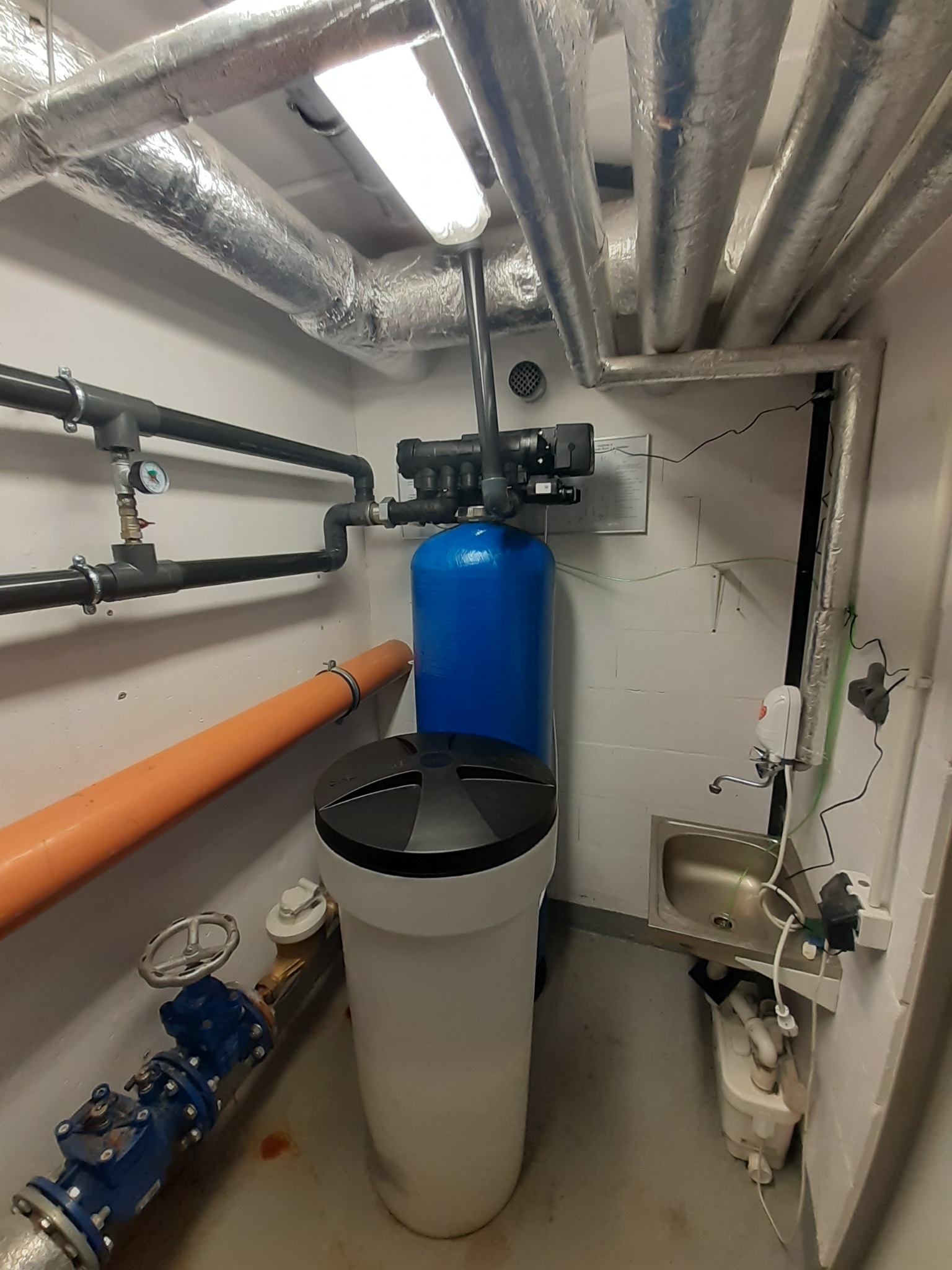 Instalacja uzdatniania wody w pomieszczeniu technicznym: niebieski zbiornik z czarną głowicą, biały zbiornik z czarną pokrywą, rury w izolacji termicznej pod sufitem, zawory i manometr na ścianie...