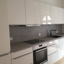 Elektro -Tech - Nowoczesna, minimalistyczna kuchnia z białymi, lakierowanymi frontami szafek, szarym blatem i płytą indukcyjną. Widoczne gniazdko elektryczne nad blatem.